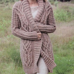 May include: A long, brown knitted cardigan with a wide, open collar and cable knit detailing. The cardigan is worn over a white dress.