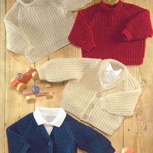Może przedstawiać: Cztery dzianinowe swetry i kardigany dla dzieci w kolorze kremowym, czerwonym i granatowym. Swetry są prążkowane i mają długie rękawy. Widoczne są również dwa drewniane samochodziki.