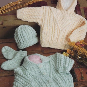 Puede incluir: Un conjunto de ropa de bebé de punto en color crema y verde claro. El conjunto incluye un jersey con capucha, un cárdigan, un gorro y manoplas.