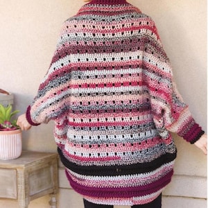 May include: A striped crochet cardigan in shades of pink, black, and gray. The cardigan has a loose fit and a wide, open neckline. The text "Sized to 3X" is visible in the upper right corner of the image.