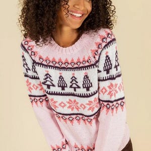May include: A light pink sweater with a crew neck and long sleeves. The sweater features a white and dark purple pattern with pink accents, including Christmas trees and snowflake designs. The sweater is made of a soft, knitted material.