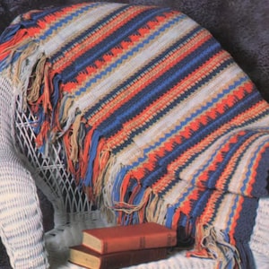 May include: A crocheted throw blanket with a striped pattern in shades of blue, red, and beige. The blanket has a fringe border and is draped over a white wicker chair.
