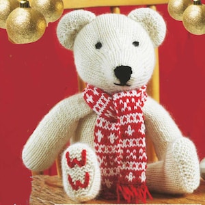 Peut inclure: Un ours en peluche blanc tricoté, orné d'une écharpe à motifs rouges et blancs. L'ours a des yeux noirs en boutons et un nez noir. Il est assis sur une chaise en bois. Le pied de l'ours porte le chiffre 33 tricoté.
