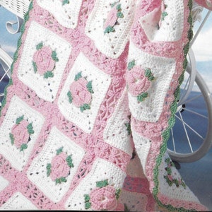 May include: A crocheted baby blanket with a pink and white square pattern and green accents. The blanket features a floral design with pink roses and green leaves.