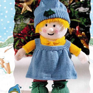Puede incluir: Muñeca de punto con un gorro azul, una camisa amarilla, un vestido azul y zapatos verdes. La muñeca está de pie frente a un fondo nevado con un árbol de Navidad.