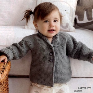 Könnte beinhalten: Graue, gestrickte Babyjacke mit Knopfverschluss. Die Jacke hat lange Ärmel und einen Kragen. Das Baby trägt die Jacke und steht vor einem weißen Hintergrund. Die Jacke ist aus weichem, strukturiertem Garn gefertigt.