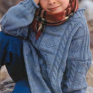 May include: A young child wearing a blue knitted sweater with a cable knit pattern and a red and black plaid scarf. The child is sitting on a rock and has their hand on their chin.