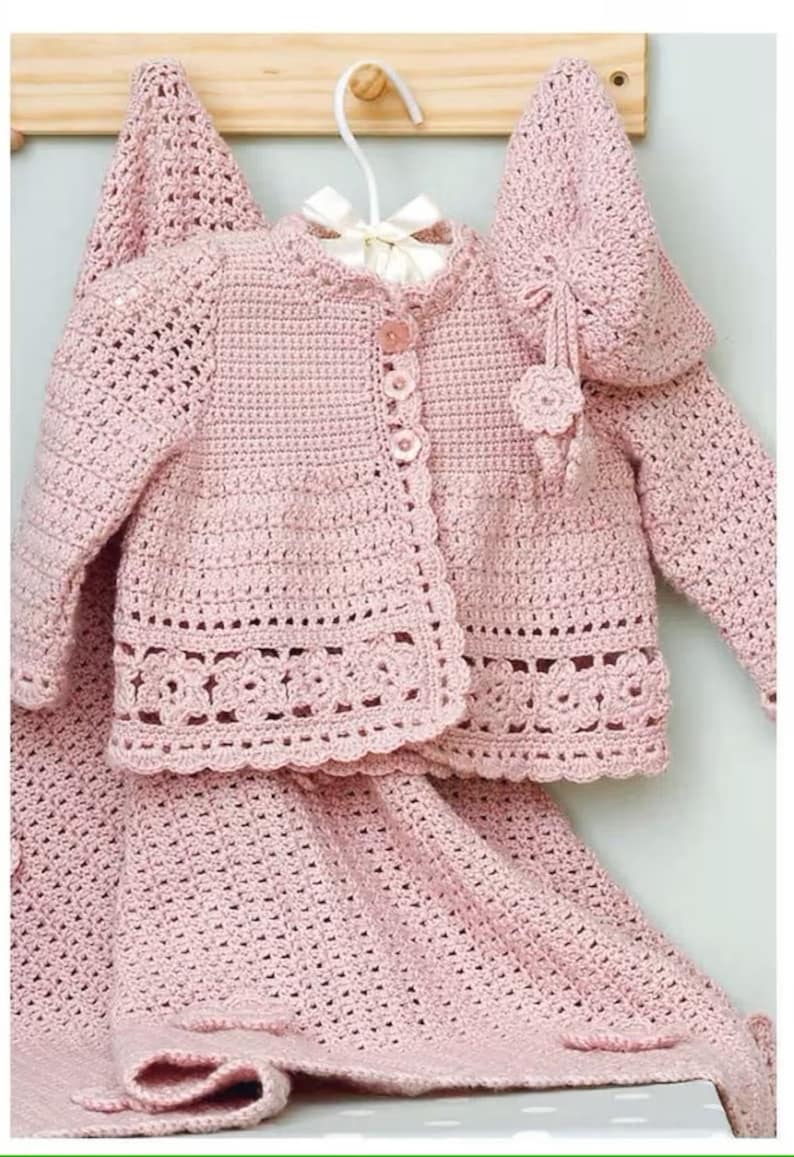 May include: A pink crocheted baby outfit set with a cardigan, skirt, and hat. The cardigan has a button closure and a delicate lace trim. The skirt is a simple crochet design. The hat has a flower embellishment.
