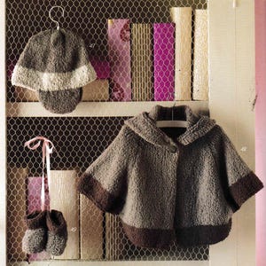 May include: A set of knitted baby clothes in brown and white. The set includes a hat, a hooded cape, and booties. The clothes are hanging on a wire rack.