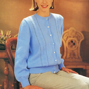 Puede incluir: Un cárdigan azul claro con cuello redondo y cierre de botones. El cárdigan presenta un diseño de rombos y mangas largas. Los botones son blancos. La persona lleva pantalones estampados y está sentada en una silla de madera.