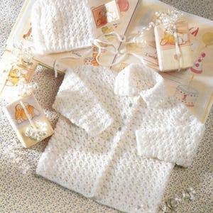 Peut inclure: Un cardigan blanc en crochet pour bébé avec un bonnet assorti. Le cardigan a une fermeture à boutons et un col. Le bonnet a un sommet pointu.