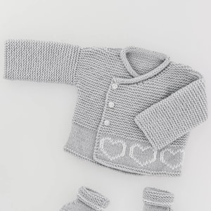 Puede incluir: Un cárdigan gris de punto para bebé con un patrón de corazón blanco en la parte inferior y cierre de botón. El cárdigan se combina con botines de punto grises a juego.