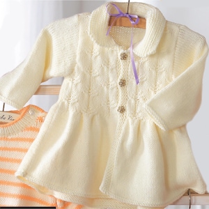 May include: Cream-colored knitted baby cardigan with a Peter Pan collar, long sleeves, and a flared skirt. The cardigan features a textured pattern on the bodice and is fastened with three flower-shaped buttons. A purple ribbon is attached to the hanger.