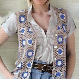 May include: A crocheted vest with a beige background and blue and white floral accents. The vest has a V-neck and buttons down the front.