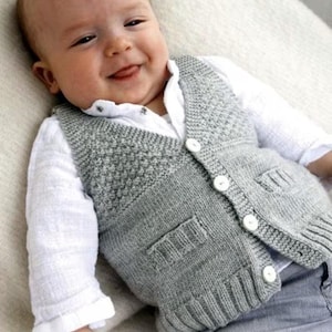 May include: A grey knitted baby waistcoat with white buttons and two front pockets. The waistcoat is worn over a white collared shirt. The baby is smiling and wearing the waistcoat.
