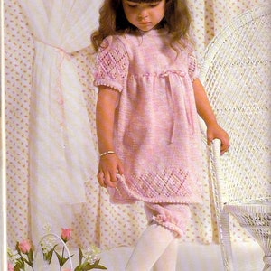 May include: A young girl wearing a pink knitted dress with a white lace trim and white tights. She is standing next to a white wicker chair. The dress has a bow at the waist and a lace pattern on the sleeves. The image is captioned "No. T95".
