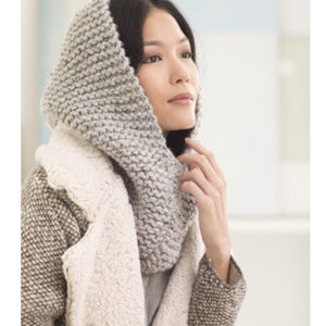 May include: A gray knitted cowl neck scarf with a textured stitch pattern. The scarf is worn by a person with dark hair and is styled to cover the head and neck.