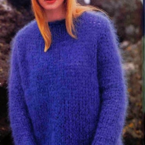 May include: A woman wearing a blue fuzzy jumper and a blue knitted hat. The jumper is a loose fit and has a turtleneck.