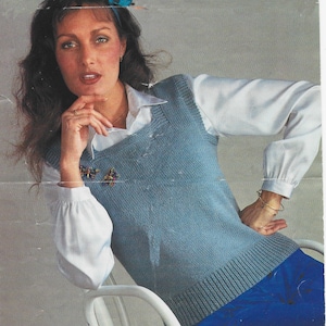 May include: A pale blue knitted vest with a ribbed hem and a butterfly brooch. The vest is worn over a white collared shirt with long sleeves. The model is wearing blue trousers.