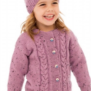 May include: A young girl wearing a purple knitted cardigan with a cable knit pattern and buttons. She is also wearing a purple knitted hat and a navy blue skirt with white polka dots.