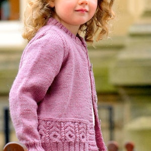 May include: A young girl wearing a pink knitted cardigan with a cable knit pattern on the bottom. The cardigan has a button closure and is worn over a white and pink striped shirt.