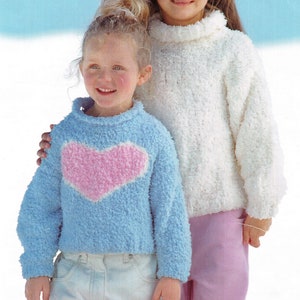 Puede incluir: Dos chicas con suéteres de punto peludos. La chica de la izquierda lleva un suéter azul claro con un corazón rosa en la parte delantera. La chica de la derecha lleva un suéter blanco.