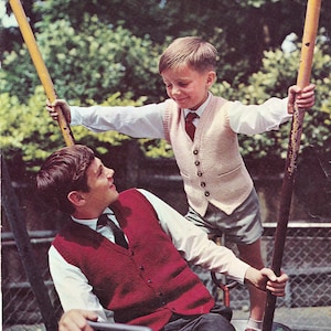May include: Two boys wearing knitted waistcoats. The boy on the left is wearing a red waistcoat and a white shirt with a black tie. The boy on the right is wearing a light pink waistcoat and a white shirt with a red tie. They are both holding onto ropes attached to a swing set.