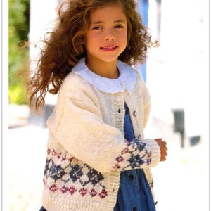Può includere: Una bambina indossa un cardigan di lana bianco con un bordo a motivi blu e rossi. Il cardigan ha una chiusura con bottoni e un colletto bianco.