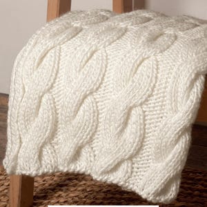 Puede incluir: Una manta de punto de cable de color crema, sobre una silla de madera. La manta presenta un tejido grueso y texturizado con un patrón repetitivo de cables entrelazados. La suave y acogedora manta es un artículo de decoración del hogar.