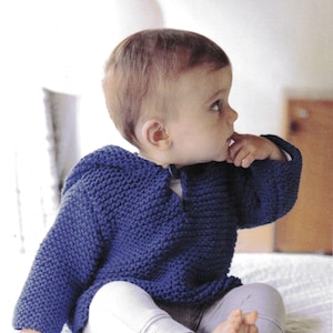 May include: A knitted blue baby sweater with a hood. The sweater is a solid, deep blue color and has a textured appearance. The baby is wearing light gray pants. The sweater appears to be handmade.