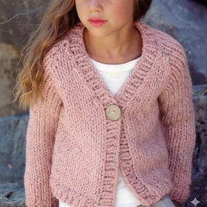 May include: A light pink knitted cardigan with a large wooden button closure. The sweater has a V-neck and a textured knit pattern. The cardigan is a soft, muted pink color, perfect for layering.