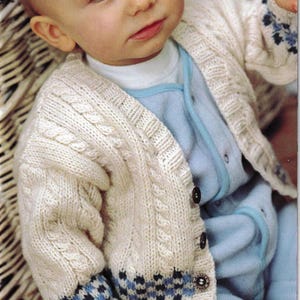 Puede incluir: Cárdigan de bebé de punto de cable color crema con puños estampados en azul y negro. El suéter tiene botones marrones oscuros y se usa sobre un mono azul claro. El cárdigan presenta un diseño de punto texturizado.
