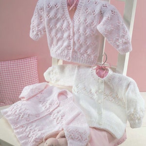 Puede incluir: Dos cárdigans de punto para bebé, uno rosa y otro blanco, con un delicado patrón de encaje. Los cárdigans son abotonados y tienen mangas largas.