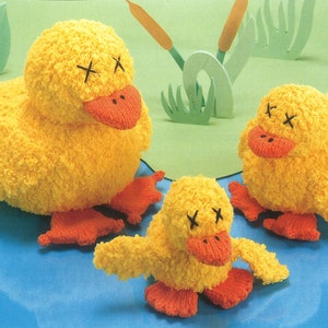 Peut inclure: Trois jouets en peluche de canard jaunes tricotés avec des becs rouges et des yeux noirs. Les canards sont couchés sur une surface bleue avec de l'herbe verte et des roseaux en arrière-plan.
