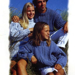 Puede incluir: Una familia de tres personas con jerséis de punto azules a juego. Los jerséis tienen un patrón texturizado y mangas largas. El fondo presenta hierba alta y un cielo azul claro, lo que sugiere un entorno al aire libre.