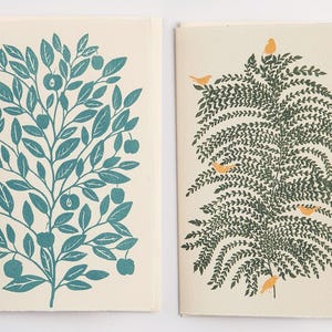 May include: Two greeting cards with nature-themed illustrations. One card features a teal tree with apples, the other has a fern-like tree with orange birds. Both cards have a cream-colored background.