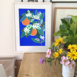 May include: A framed print of a stylized orange tree with white blossoms and orange fruit on a blue background. The print is hanging on a wall above a wooden table with a vase of flowers.