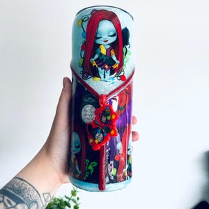 Puede incluir: Un vaso con una imagen de dibujos animados de Sally de The Nightmare Before Christmas. El vaso tiene una base de purpurina azul claro con un dise&ntilde;o de cremallera roja. La imagen de Sally est&aacute; rodeada de luces navide&ntilde;as.