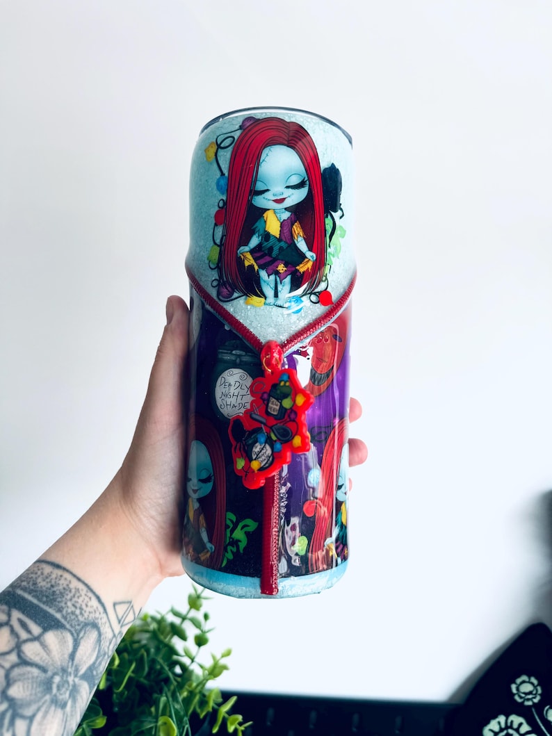 Puede incluir: Un vaso con una imagen de dibujos animados de Sally de The Nightmare Before Christmas. El vaso tiene una base de purpurina azul claro con un dise&ntilde;o de cremallera roja. La imagen de Sally est&aacute; rodeada de luces navide&ntilde;as.