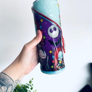 Puede incluir: Un vaso cil&iacute;ndrico con personajes de Pesadilla antes de Navidad. El vaso tiene un fondo morado con ilustraciones de personajes, una parte superior azul claro y un dise&ntilde;o de cremallera roja. El vaso est&aacute; sostenido por una persona.