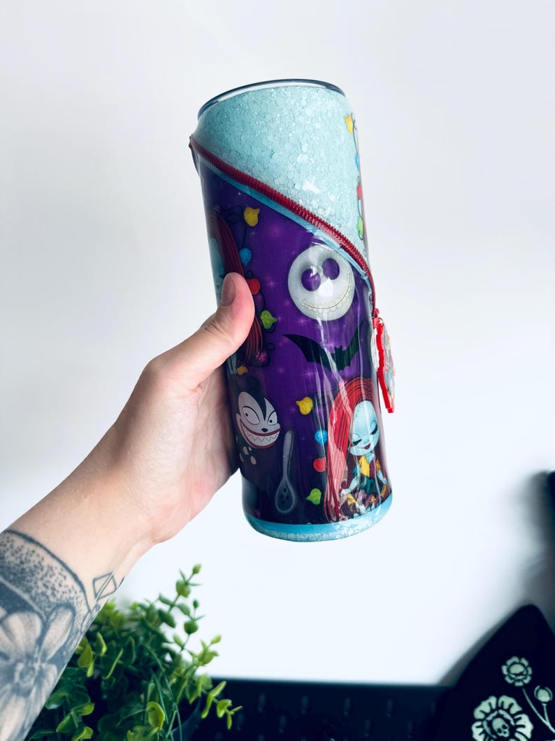 Puede incluir: Un vaso cil&iacute;ndrico con personajes de Pesadilla antes de Navidad. El vaso tiene un fondo morado con ilustraciones de personajes, una parte superior azul claro y un dise&ntilde;o de cremallera roja. El vaso est&aacute; sostenido por una persona.