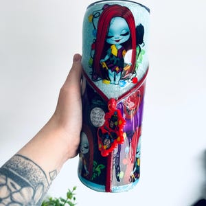 Puede incluir: Un vaso con un personaje de dibujos animados con cabello rojo y tez azul, adornado con luces navide&ntilde;as. El dise&ntilde;o incluye un detalle de cremallera y el texto "Deadly Night Shade". El vaso es sostenido por una persona.