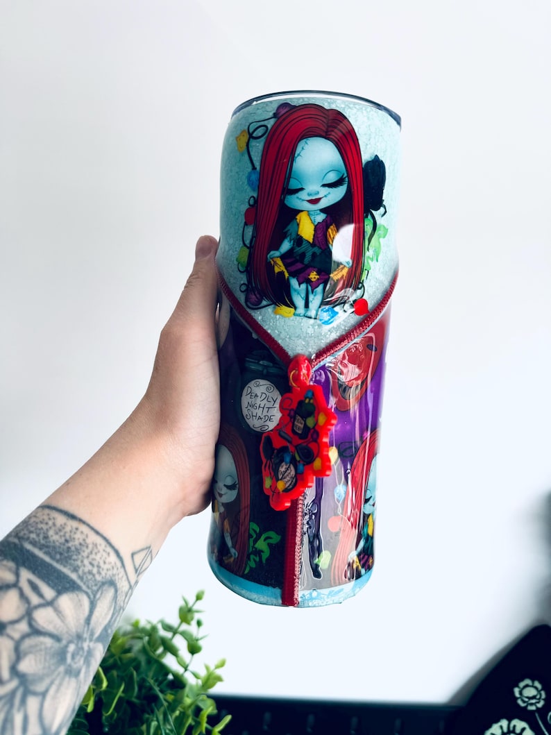 Puede incluir: Un vaso con un personaje de dibujos animados con cabello rojo y tez azul, adornado con luces navide&ntilde;as. El dise&ntilde;o incluye un detalle de cremallera y el texto "Deadly Night Shade". El vaso es sostenido por una persona.