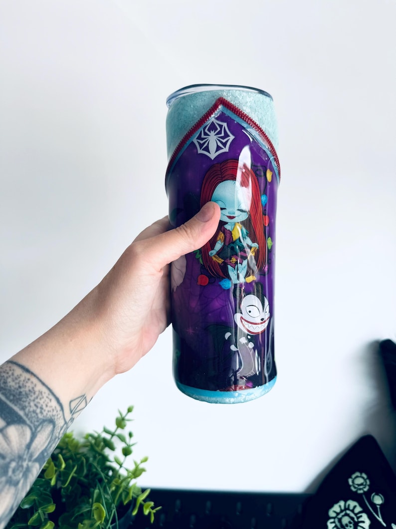 Puede incluir: Un vaso morado y azul con una imagen de dibujos animados de Sally de Pesadilla antes de Navidad. El vaso tiene un acabado brillante y un dise&ntilde;o de cremallera roja. El dise&ntilde;o incluye una telara&ntilde;a y un personaje de dibujos animados.
