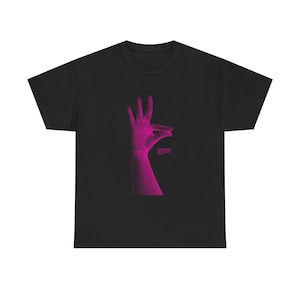 Könnte beinhalten: Schwarzes T-Shirt mit einem stilisierten, pixeligen magenta Handmotiv. Das Design zeigt eine Handgeste mit Text darunter. Das T-Shirt hat einen Rundhalsausschnitt.