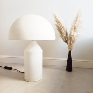 May include: A white mushroom-shaped table lamp with a white cord and a white base. The lamp is on a light gray floor. A vase with dried pampas grass is in the background.