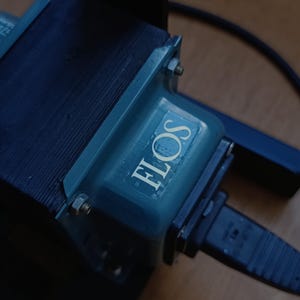 May include: A close-up of a blue electrical transformer with the word "FLOS" printed on it. The transformer is connected to a black electrical cord with a plug.