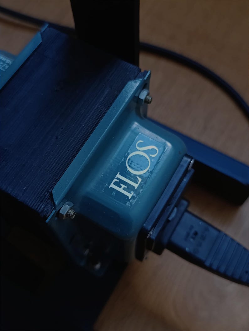 May include: A close-up of a blue electrical transformer with the word "FLOS" printed on it. The transformer is connected to a black electrical cord with a plug.
