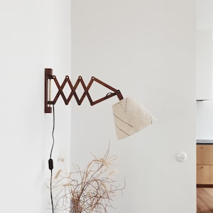 May include: A wooden wall-mounted lamp with a white fabric shade. The lamp has a folding arm design and is attached to a white wall. The lamp is turned on and the light is shining.