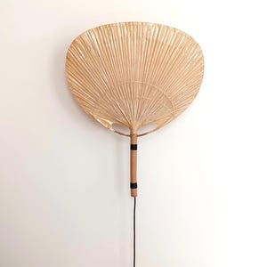 May include: A wall-mounted light fixture shaped like a large, natural, woven fan. The fan is made of light brown, dried palm leaves and has a wooden handle with black tape accents. The light fixture has a black cord and a black plug.
